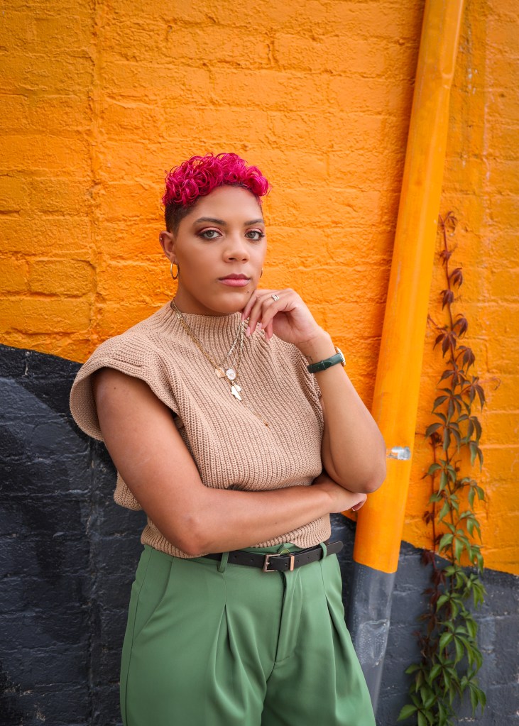 Sarah is looking contemplative in a head shot. Her hair is died pink, and she is wearing a simple brown sweater vest with a bright orange brick background. 