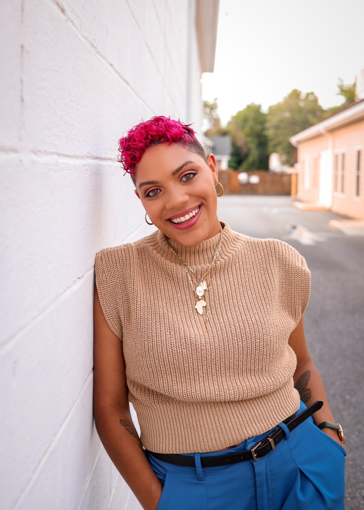 Sarah is smiling in a head shot. Her hair is died pink, and she is wearing a simple brown sweater vest with a white brick background. 