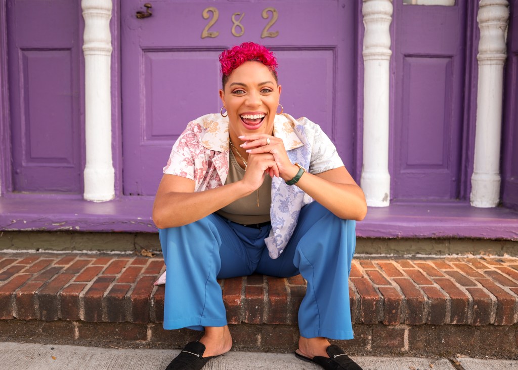 Sarah is smiling in a picture sitting in a colorful outfit in front of a purple door